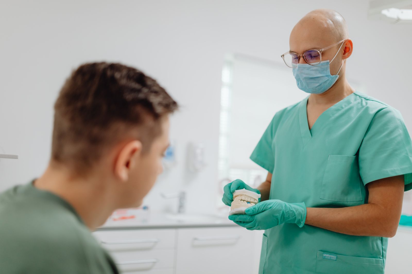 A dentist in scrubs showing a model of a jaw to a patient across from them in a treatment room.